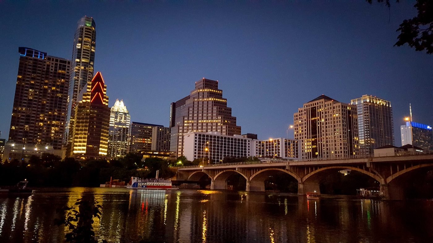 Downtown Austin at dusk, seen across Lady Bird Lake. The skyline is lit against a deep blue sky and the Congress Avenue bridge crosses the water on the right.