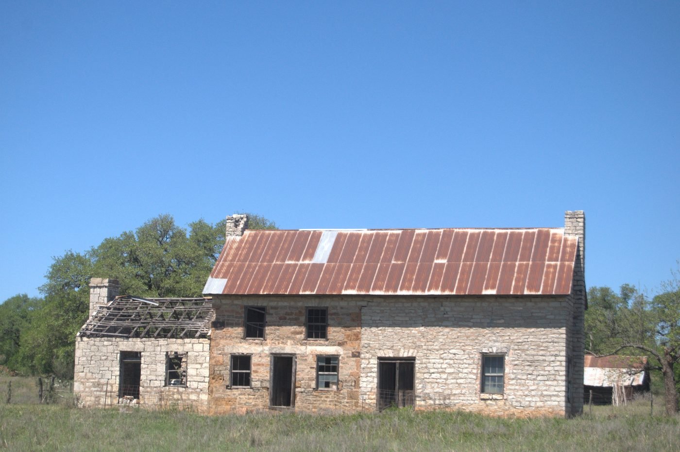 An abandoned limestone farmhouse in the Texas hill country. The cut-stone walls are intact, the rusted tin roof is failing, and the windows are empty, under a clear blue sky.